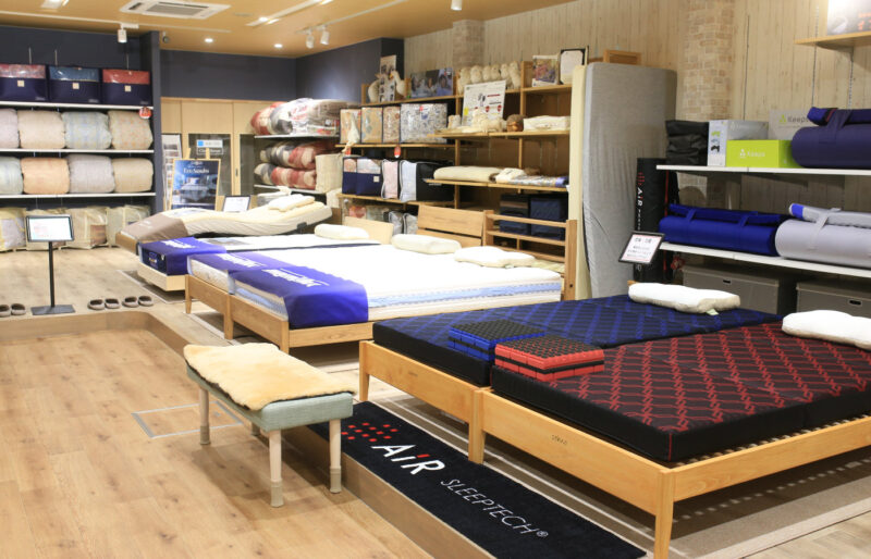 寝具と枕の専門店もりたけの店内風景。手前には「西川エアー(AiR)」のマットレス体験コーナーがあり、奥に向かって複数のベッドマットレスが設置され、実際に寝比べができる広々とした空間です。壁際の棚には羽毛ふとんなどの寝具類が豊富に陳列されています。