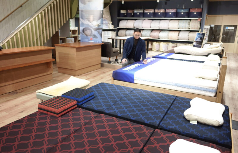 寝具と枕の専門店もりたけの店内風景。手前の「西川エアー(AiR)」などのマットレス体験コーナーで、専門スタッフがベッドマットレスの案内やフィッティングの準備をしている様子。奥には種類豊富な寝具が陳列されています。