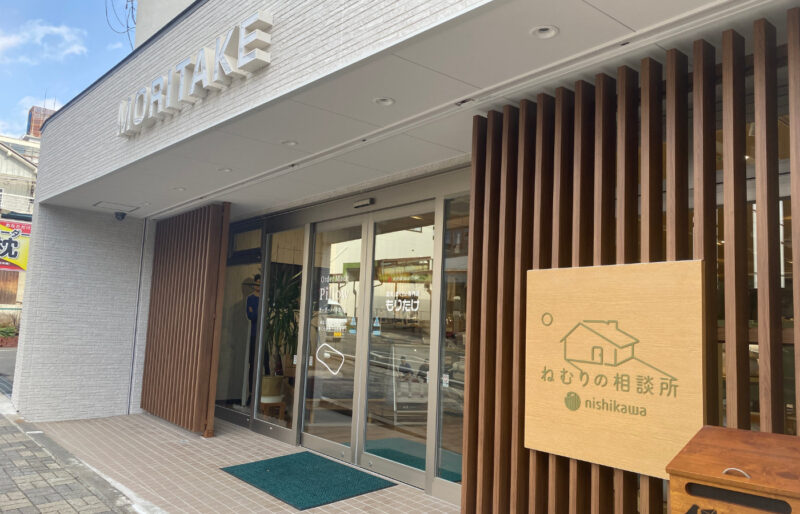 寝具と枕の専門店もりたけの店舗外観。白を基調としたモダンな建物の入り口には「MORITAKE」の立体ロゴが掲げられ、手前の木格子には西川「ねむりの相談所」の木製看板が設置されています。