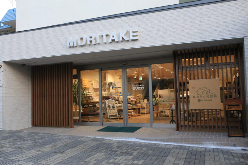 寝具と枕の専門店もりたけの店舗正面の外観写真。白い外壁に「MORITAKE」のロゴが掲げられ、右側の木格子には西川「ねむりの相談所」の看板が設置されています。ガラス張りの入り口からは、豊富な寝具が並ぶ明るい店内の様子が確認できます。