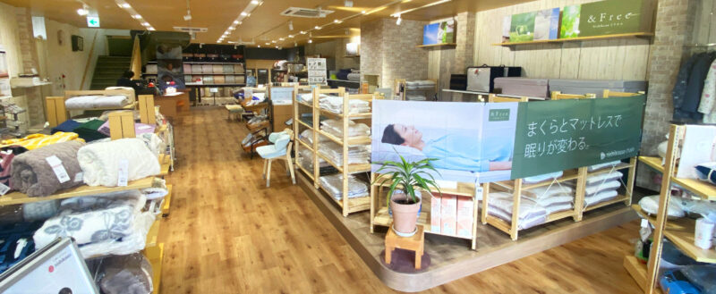 寝具と枕の専門店もりたけの店内風景。温かみのある木目調のフロアに、毛布や敷きパッドなどの寝具が豊富に並んでいます。手前には西川のブランド「&Free」の特設コーナーがあり、「まくらとマットレスで眠りが変わる。」という横断幕が掲示されています。
