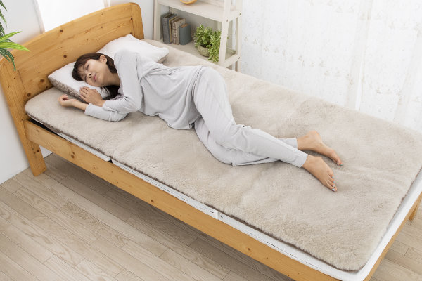 西川のムートンシーツ「シープトン」で横向きに眠る女性のイメージ画像。超高密度の毛が「天然のスプリング」となり、重みがかかりやすい肩や腰を優しくしっかりと受け止めています。人工素材では再現が難しい「究極の体圧分散」によって、ご自身の身体にしっくりと馴染む無理のない寝姿勢をサポートします。